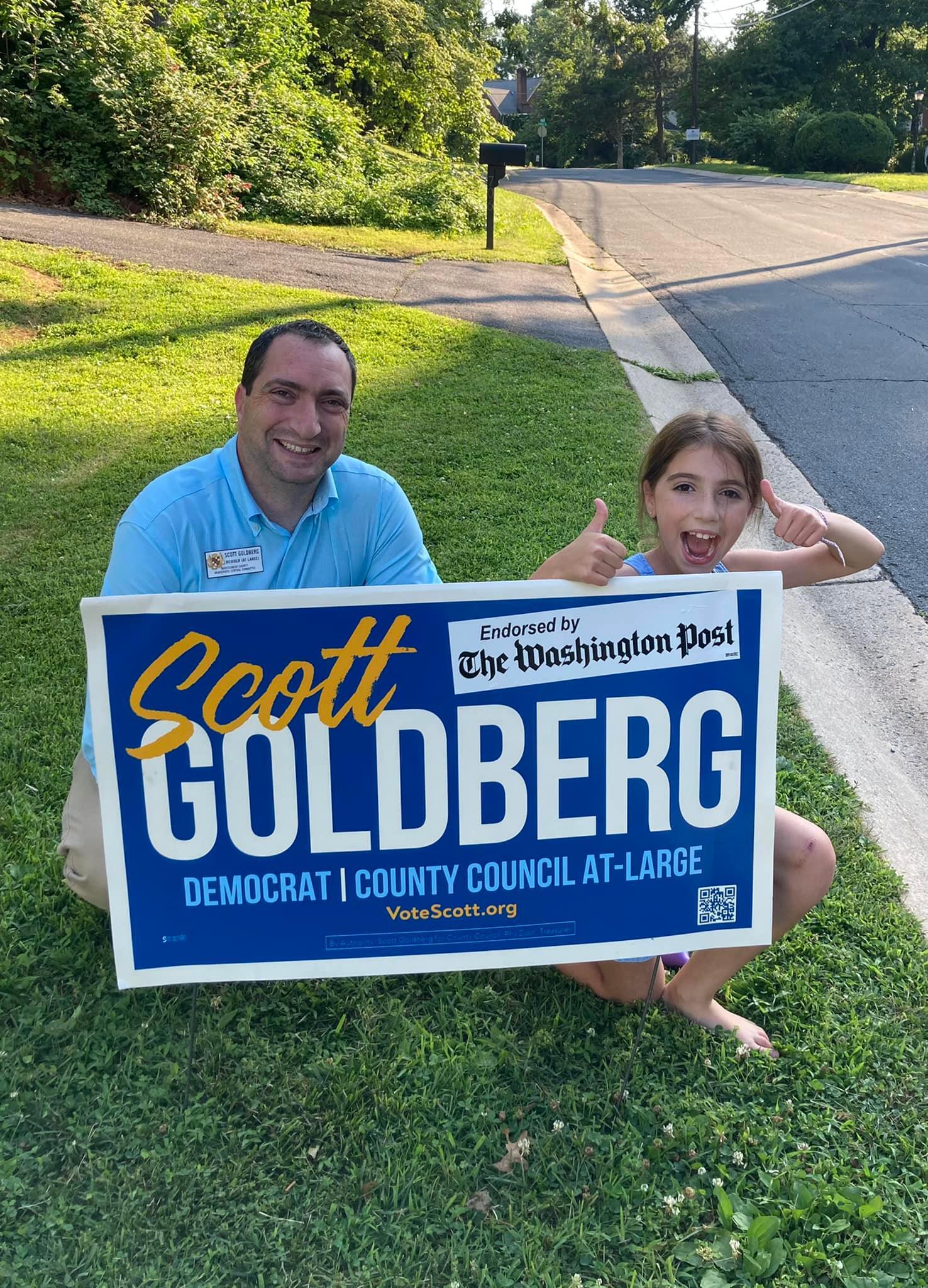 Scott with his daughter behind a campaign sign. Both are smiling and the daughter is making an enthusiastic double thumbs-up gesture.